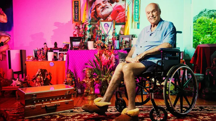 Ram Dass in wheelchair smiling with altar in background and neem karoli baba picture maharaji